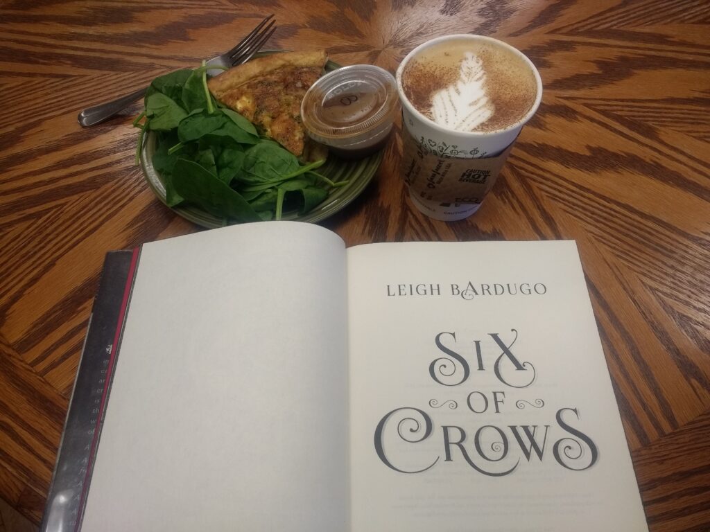 Six of Crows by Leigh Bardugo on a table beside quiche and a latte