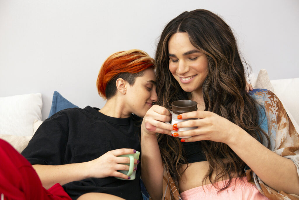 A transmasculine gender-nonconforming person and transfeminine non-binary person drinking coffee in bed