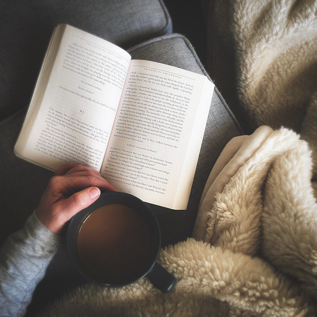 An open book next to a blanket and a person's hand holding a cup of tea